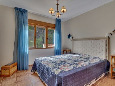 Slaapkamer met groot bed, wit gestoffeerd hoofdbord en blauw gedessineerde sprei. Terracotta tegelvloer en houten raam met blauwe gordijnen bieden uitzicht op groen.