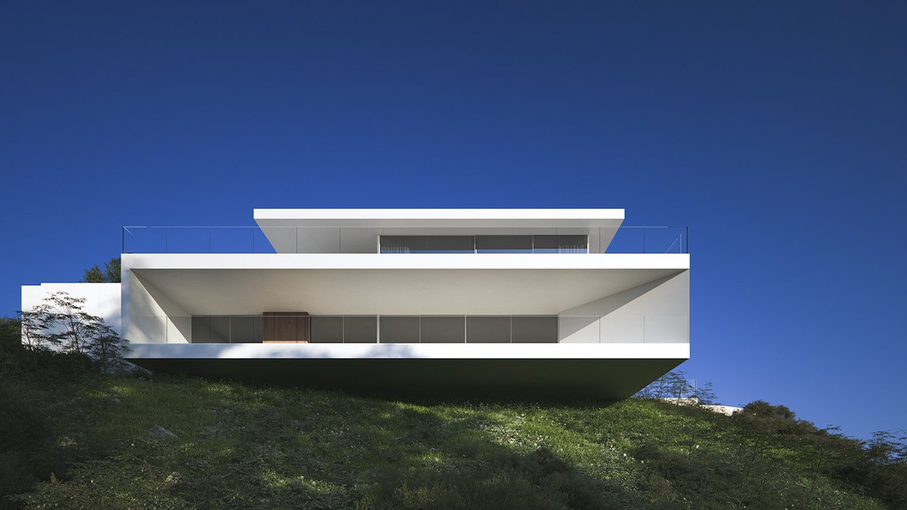 Buitengevel met minimalistische witte geometrische overstekken en glazen balustrades, geïntegreerd in een natuurlijke helling.