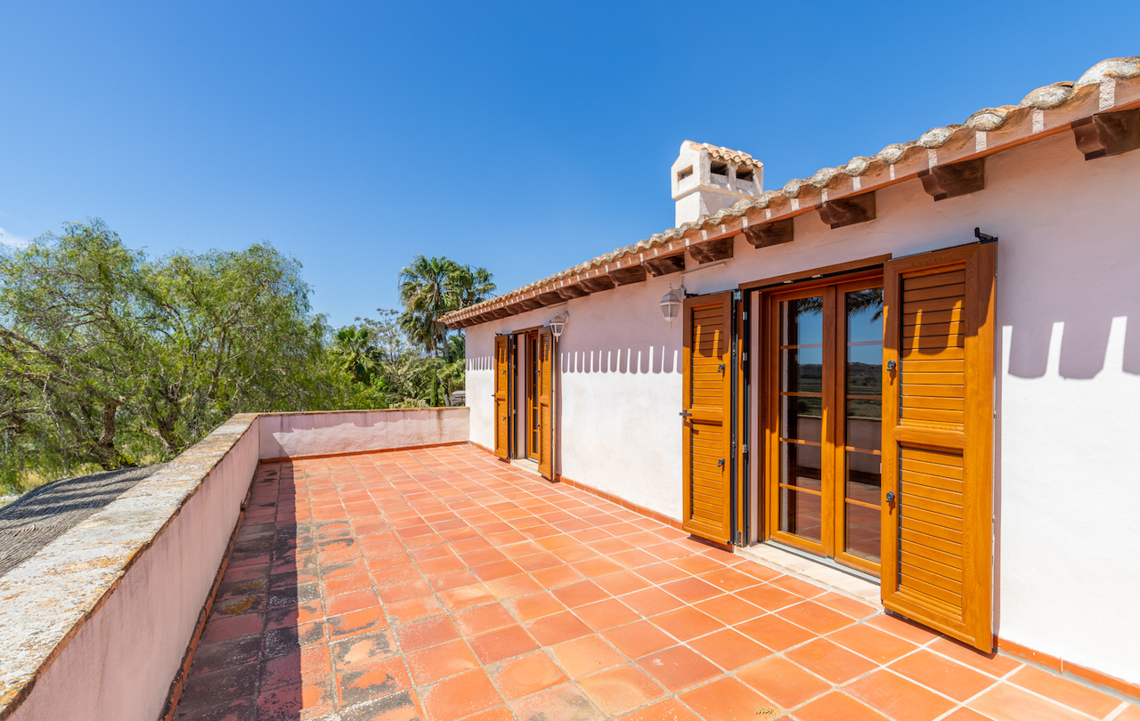 Bovenliggend terras met terracotta tegelvloer, zichtbare houten balken onder de dakrand en houten openslaande deuren met luiken tegen een witte gevel.