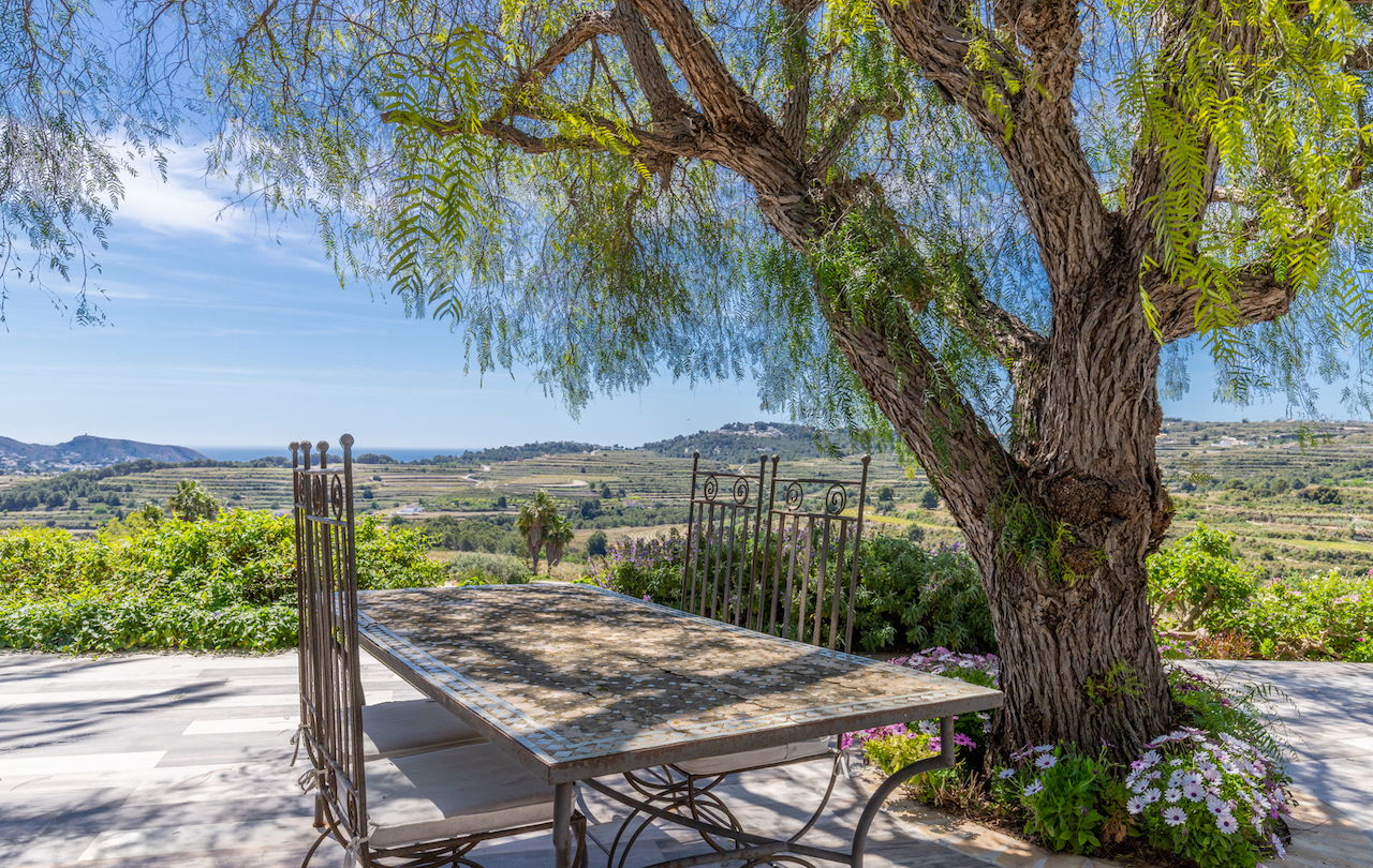 Buitenterras met een mozaïektafel onder een schaduwrijke boom, met panoramisch uitzicht over de wijngaarden en de Middellandse Zee richting Moraira.