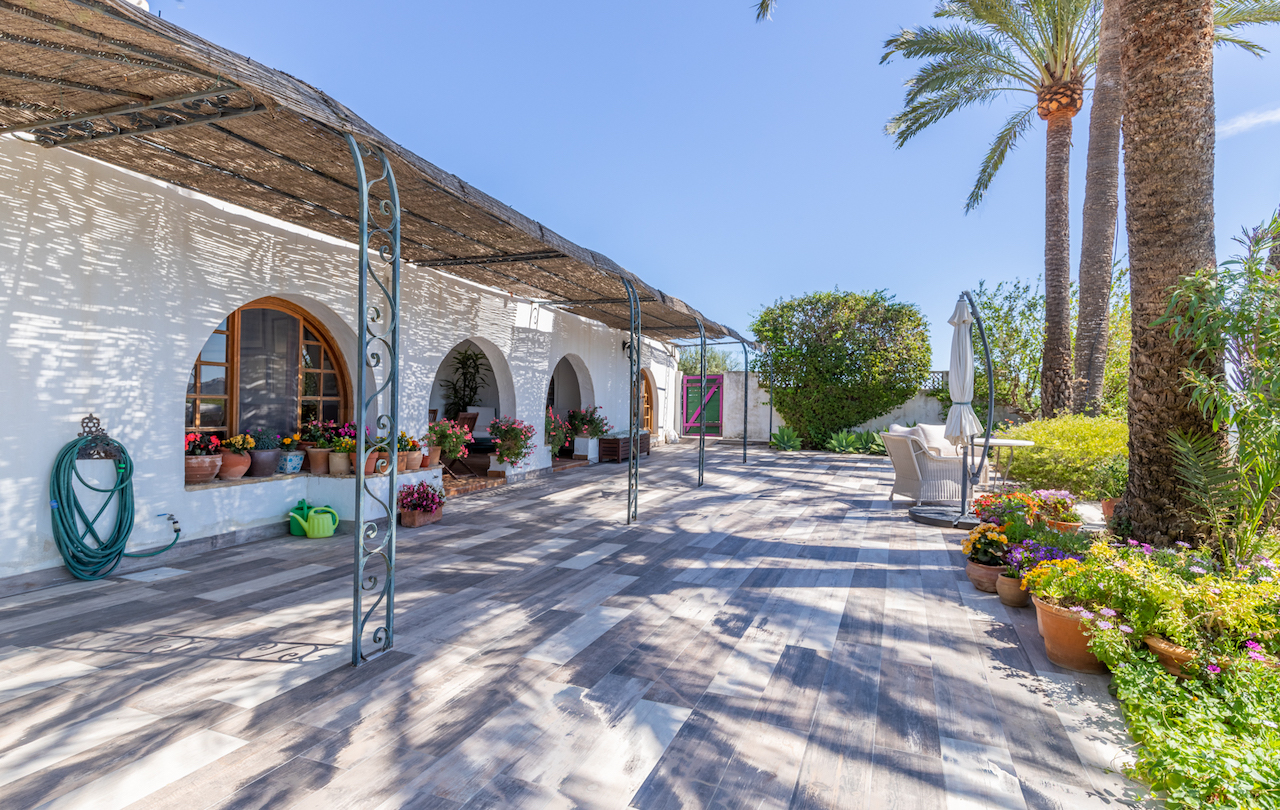 Buitenterras met houtlook tegels, een rieten pergola op smeedijzeren zuilen en een witte gevel met traditionele bogen en diverse bloempotten.