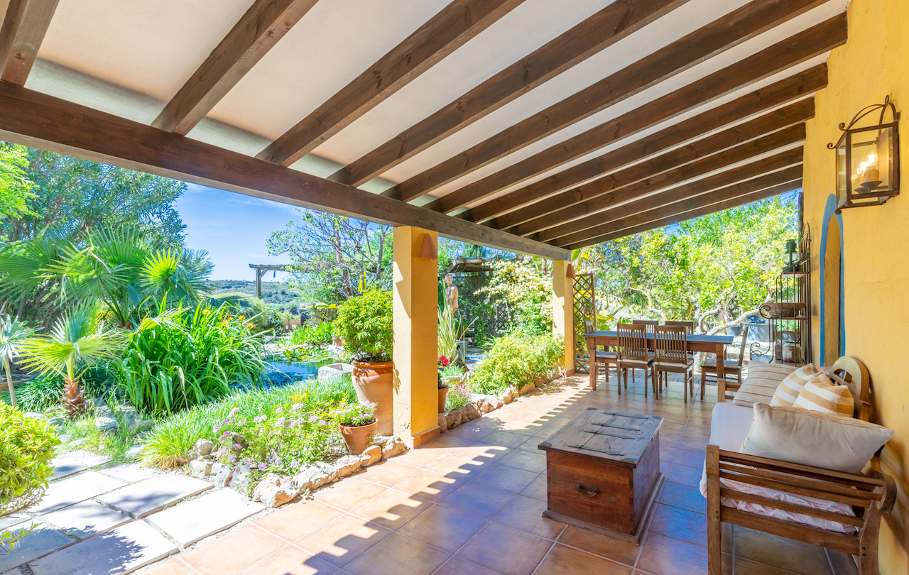 Overdekt terras met houten balkenplafond, terracotta vloer en buitenruimte met uitzicht op de volgroeide mediterrane tuin met vijver.