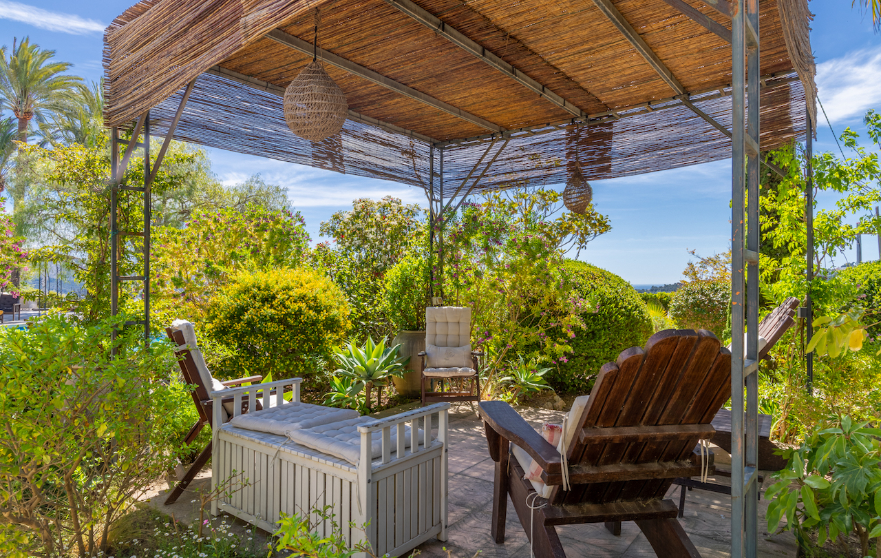 Pergola met metalen frame en rieten dak met een zithoek, omgeven door een mediterrane tuin en een weids uitzicht op de kust.
