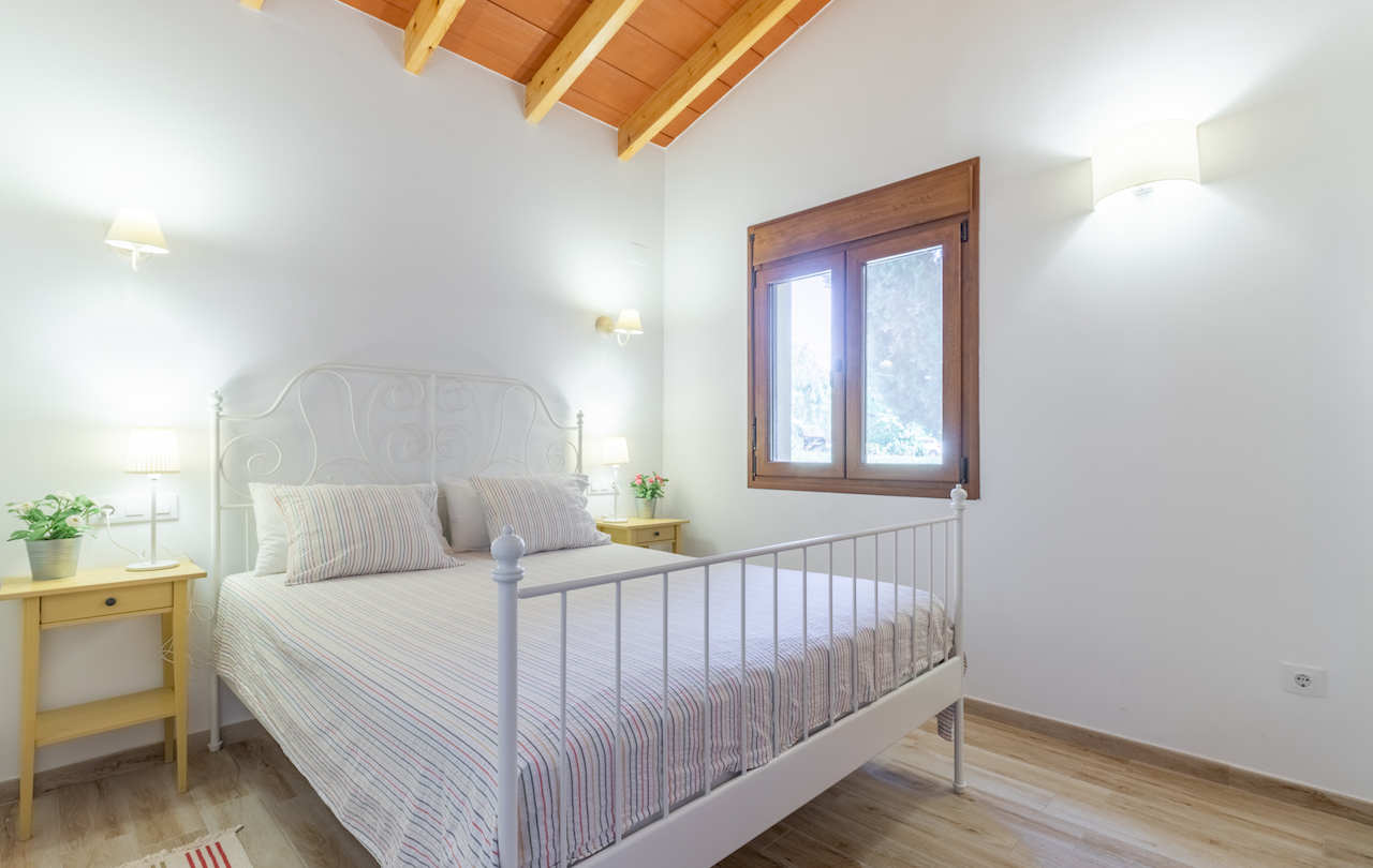 Rustieke slaapkamer met houten balkenplafond, een wit gesmeed ijzeren bed en houten kozijnen met uitzicht op de tuin.