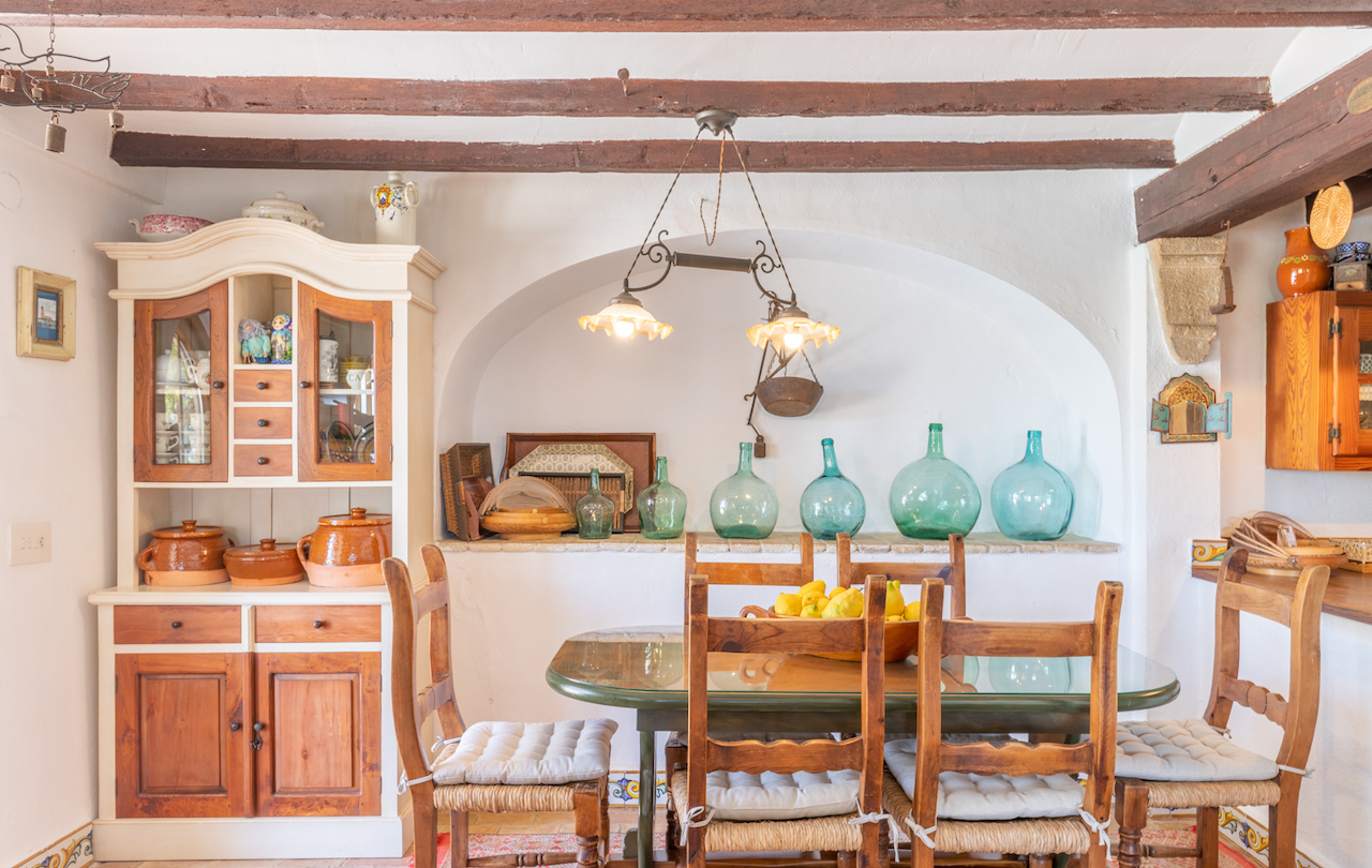 Rustieke Valenciaanse eethoek met houten balkenplafond, een traditionele servieskast en een decoratieve nis met glazen mandflessen achter de eettafel.