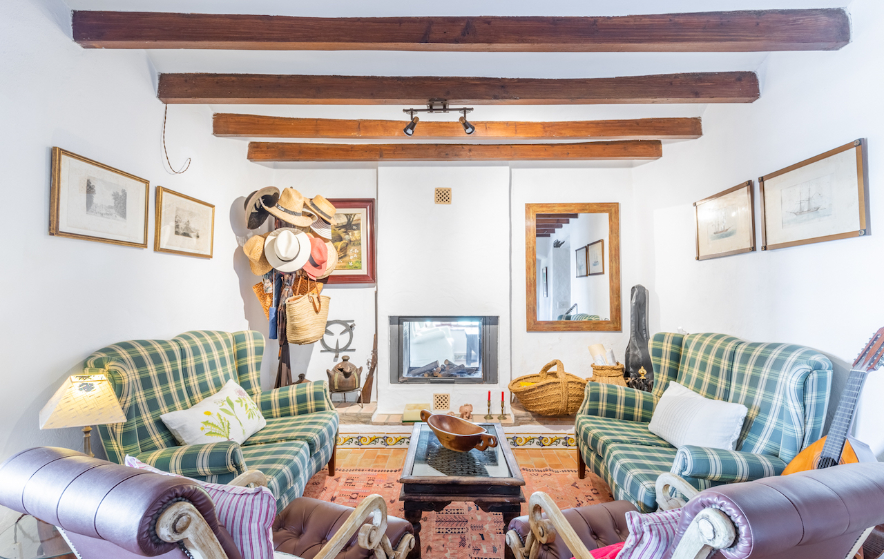 Rustieke woonkamer met donkere houten balkenplafonds, een ingebouwde haard en terracotta vloeren met decoratieve tegels. Ingericht met geruite fauteuils en traditionele details.