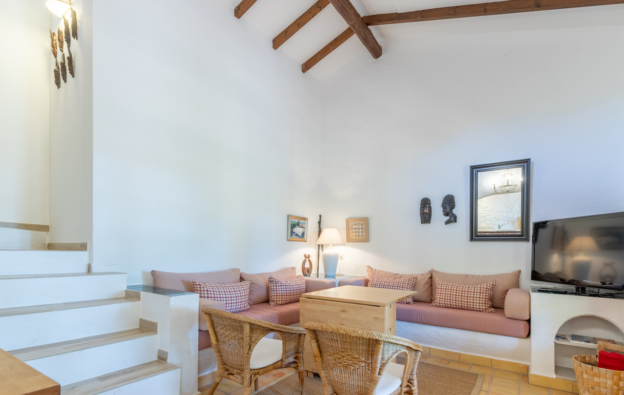 Rustieke woonkamer met hoge plafonds en houten balken. Voorzien van ingebouwde banken, rieten stoelen, een terracotta vloer en een trap met houten treden.