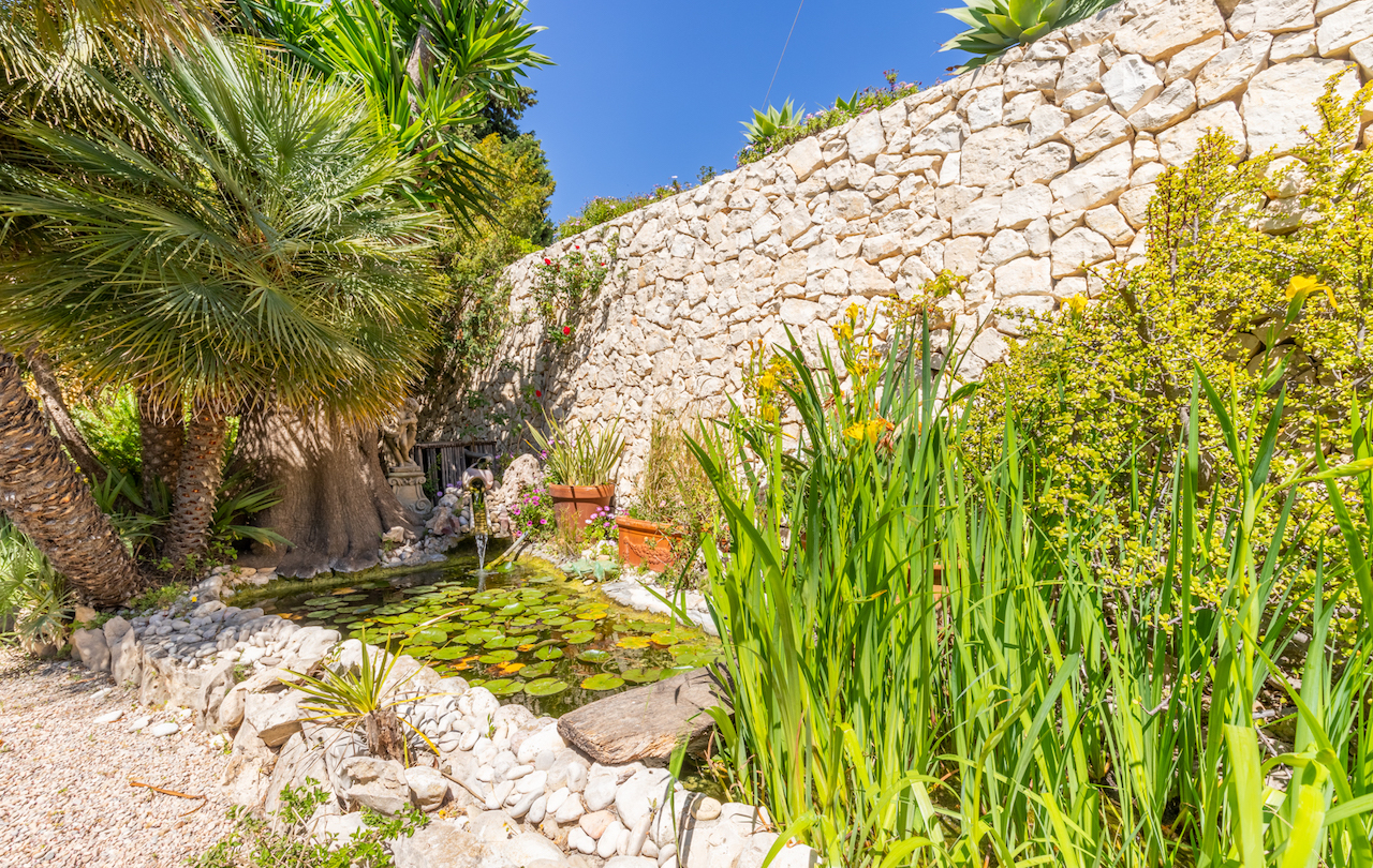 Siervijver met waterlelies en een kleine waterval, omringd door mediterrane beplanting en een traditionele droge stenen muur.