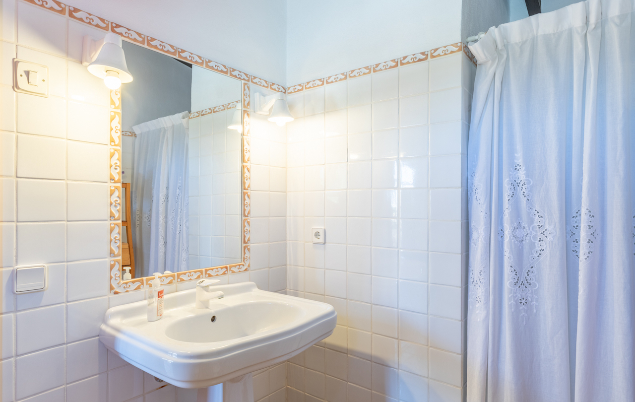 Traditionele badkamer met een witte wastafel op zuil, een spiegel met decoratieve keramische rand en klassieke wandlampen naast een geborduurd gordijn.