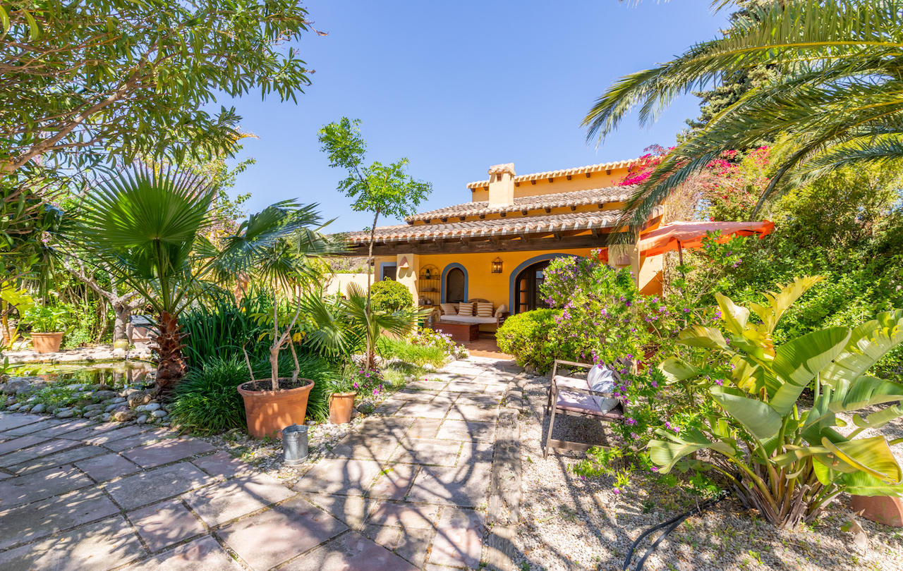 Traditionele gevel met houten balken veranda, gelegen in een mediterrane tuin met vijver, palmbomen en natuurstenen wandelpaden.