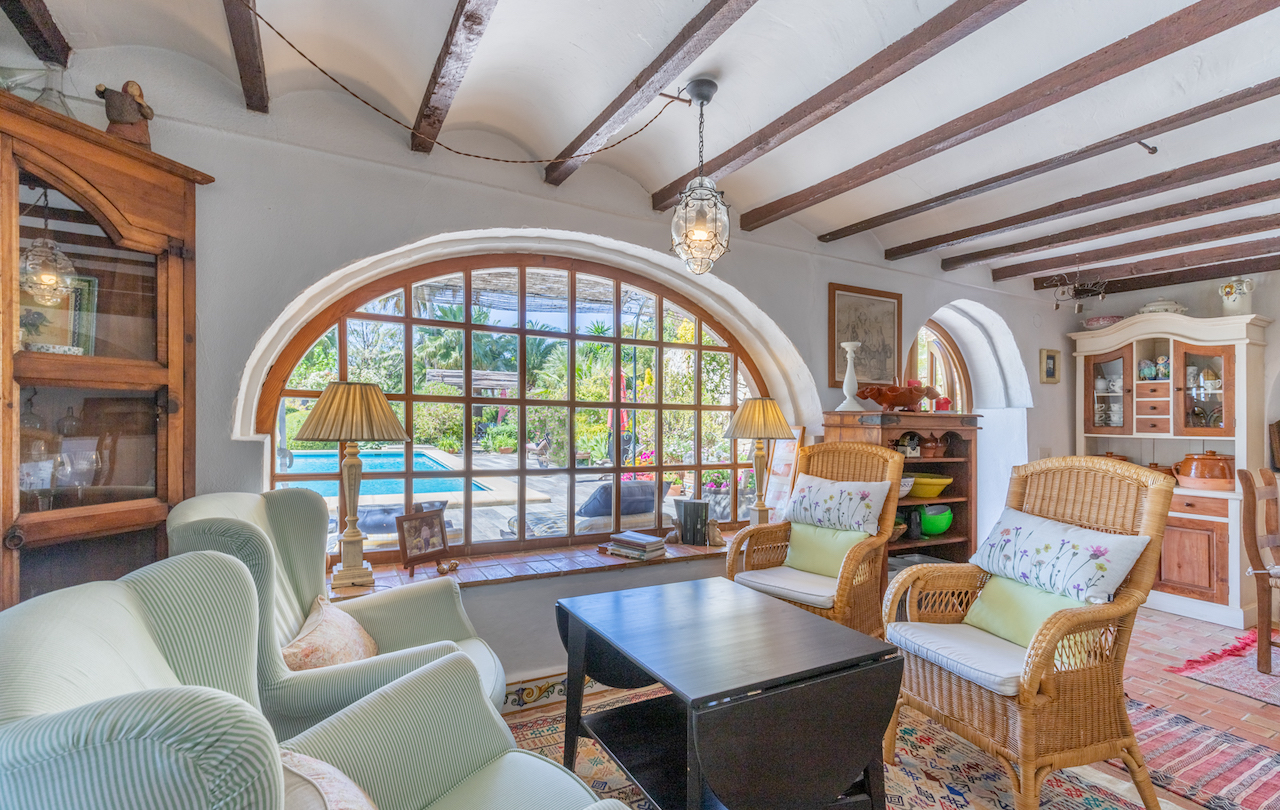 Traditionele woonkamer met houten balkenplafond, terracotta vloer en een groot boogvenster dat uitzicht biedt op het zwembad en de weelderige tuin.