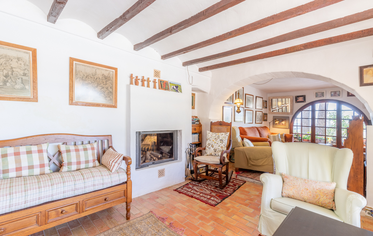 Traditionele woonkamer met houten balkenplafond, terracotta vloertegels en een inbouwhaard. Een boog verbindt de ruimte met een lichte zithoek bij een rond raam.