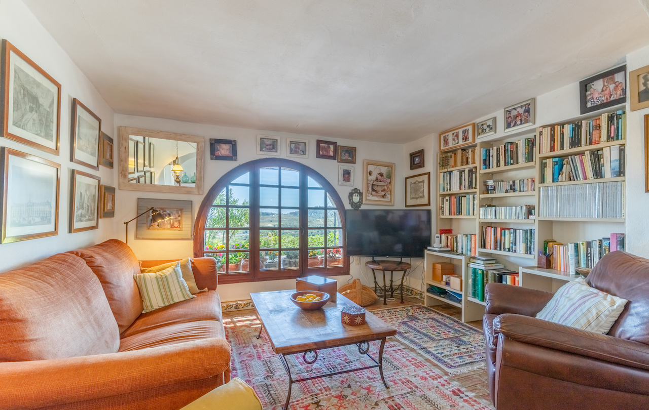 Woonkamer-bibliotheek met een groot houten boogvenster, ingebouwde boekenkasten, terracotta vloer met tapijten en vrij uitzicht op de landelijke omgeving.