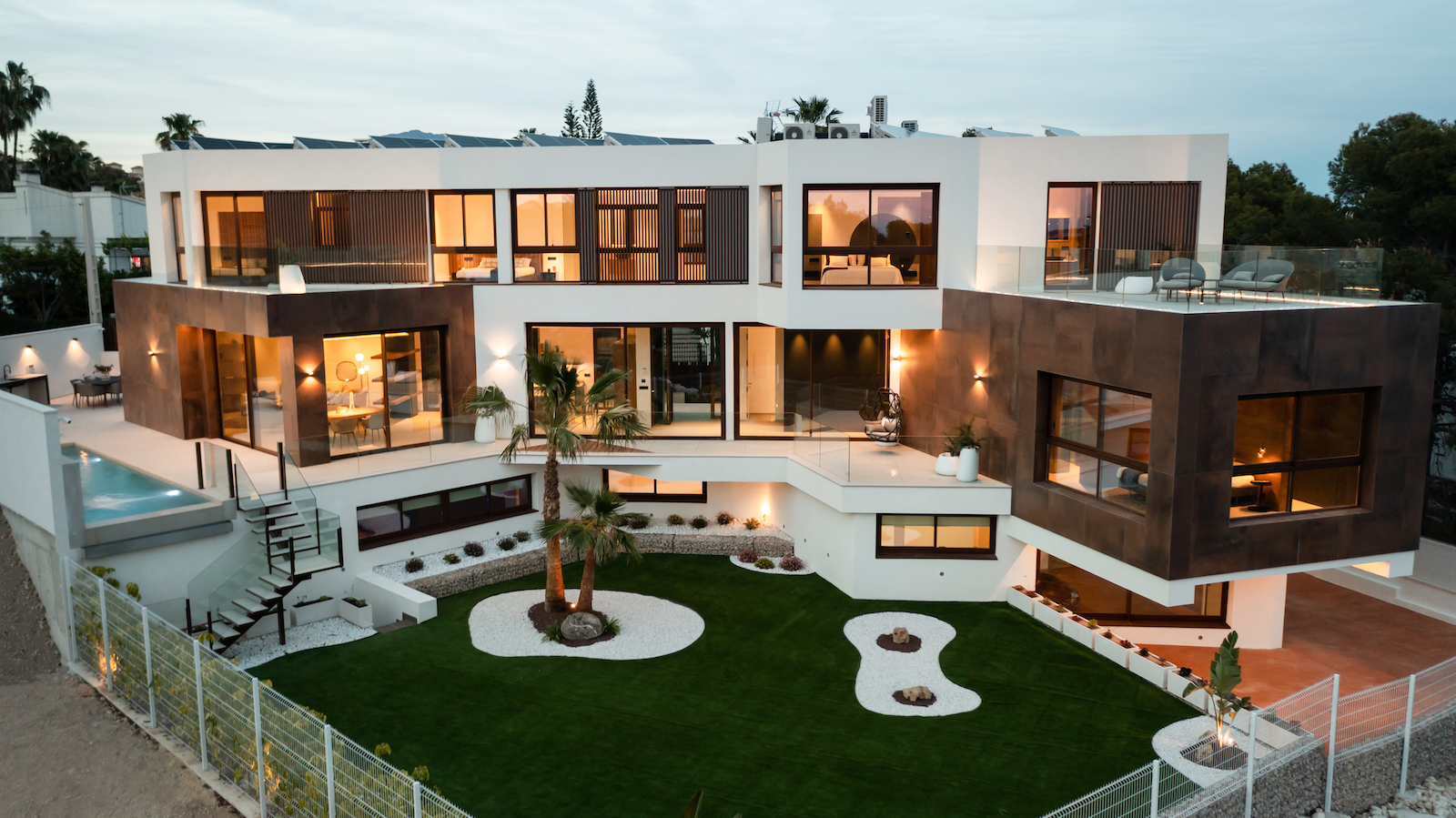Buitenaanzicht van de moderne villa met mediterrane tuin, zwembad en verlichte terrassen.