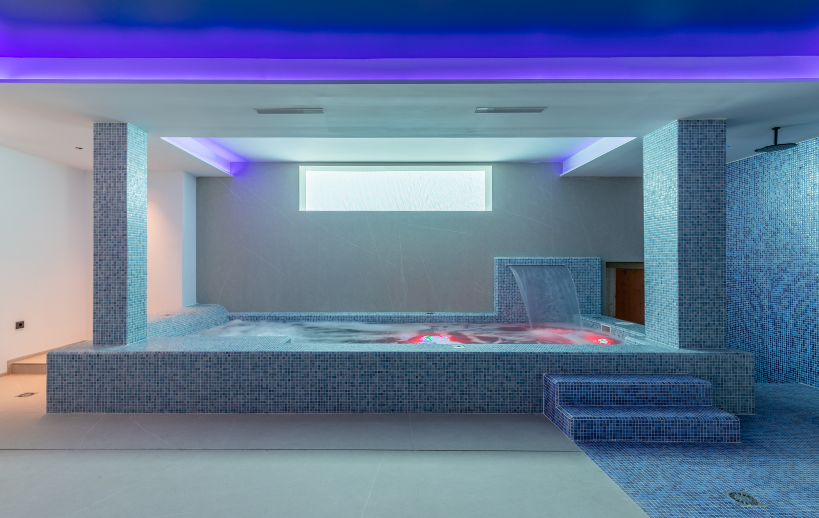 Detail van de indoor spa met jacuzzi, waterval en douche, met blauwe mozaïeken en sfeerverlichting.
