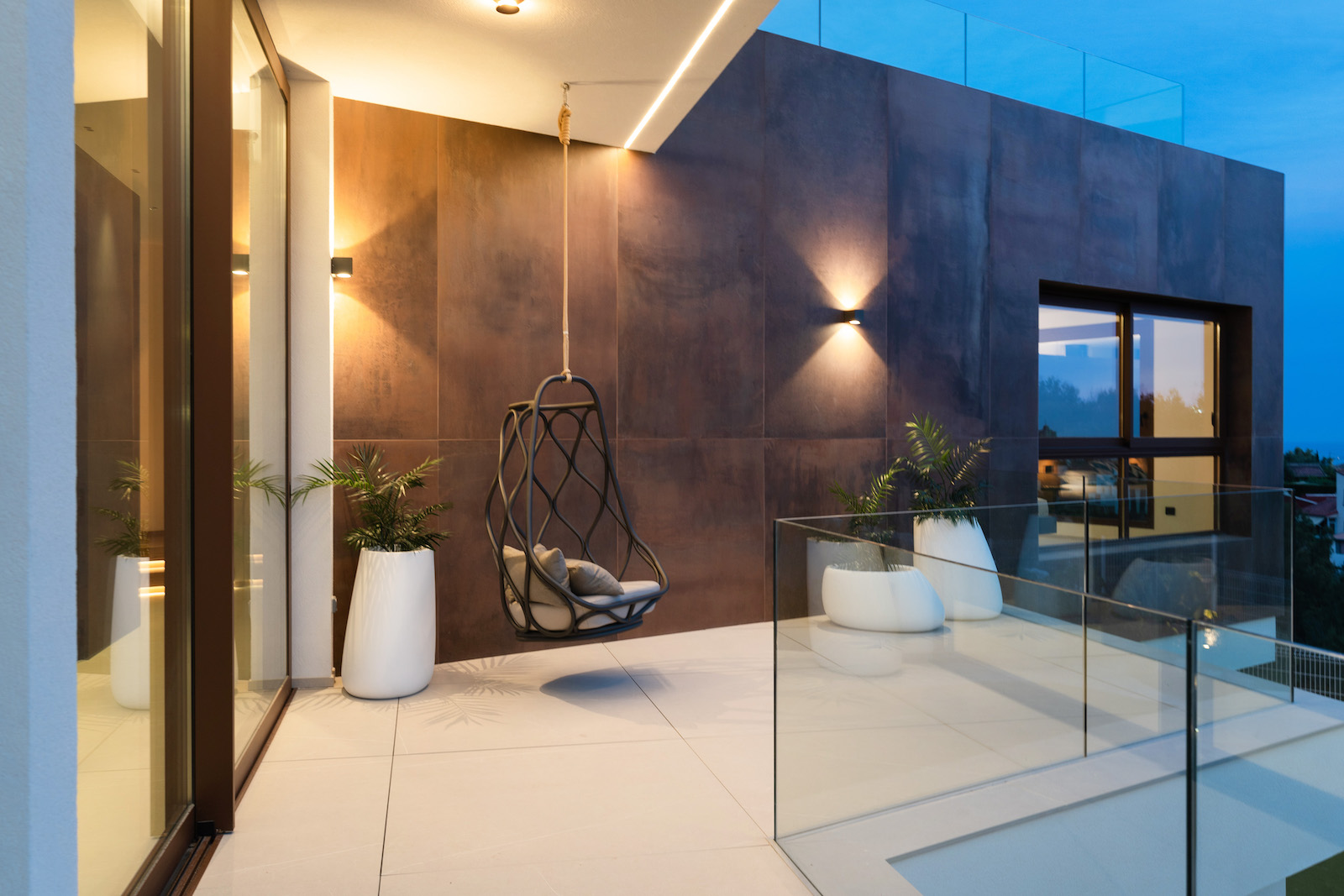 Terras met roestkleurige tegelwand, hangstoel en glazen balustrade.