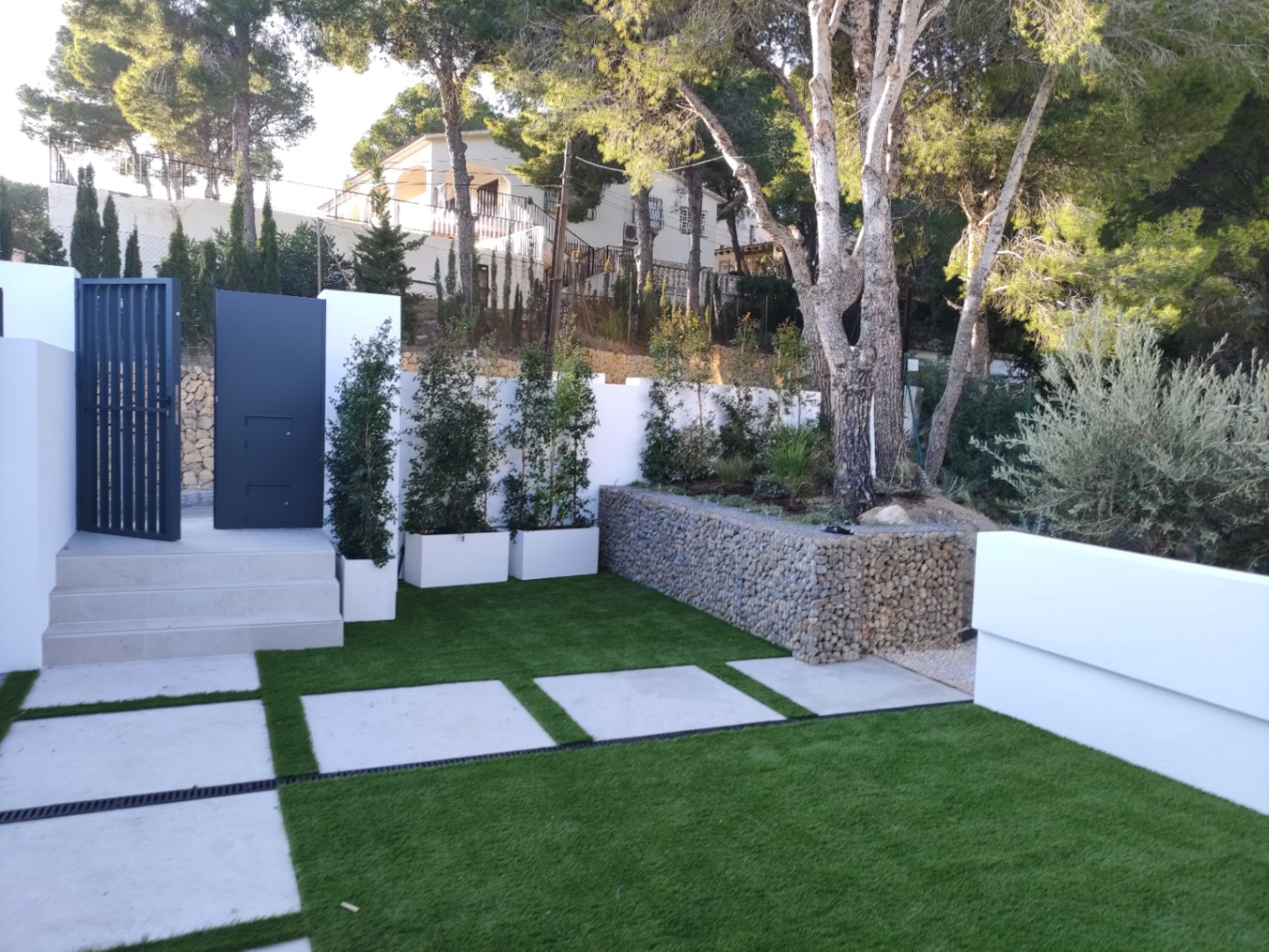 Buiteningang met modern metalen hek, stenen treden en aangelegde tuin met kunstgras, betonnen tegels en een decoratieve schanskorfmuur tussen de bomen.