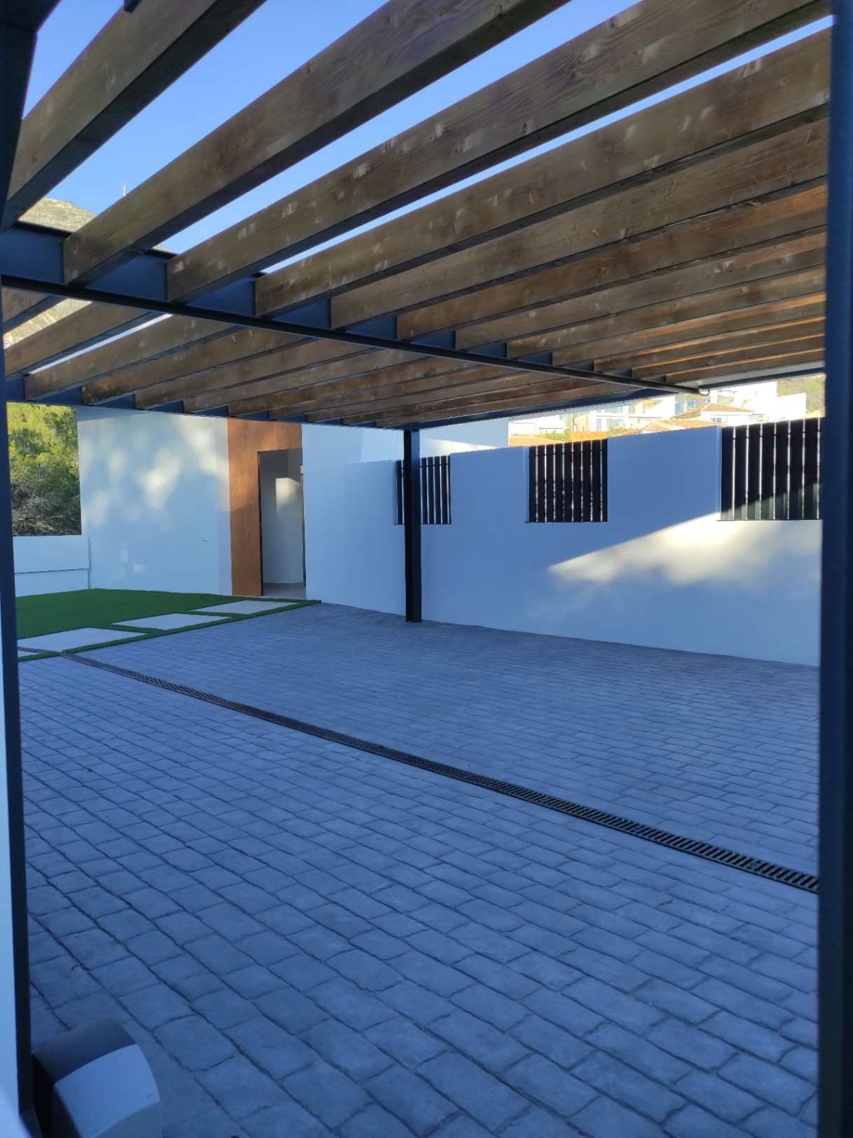 Buitenparkeerplaats met een houten balkenpergola, grijze kasseibestrating en een moderne entree met natuurlijke houten details en witte muren.