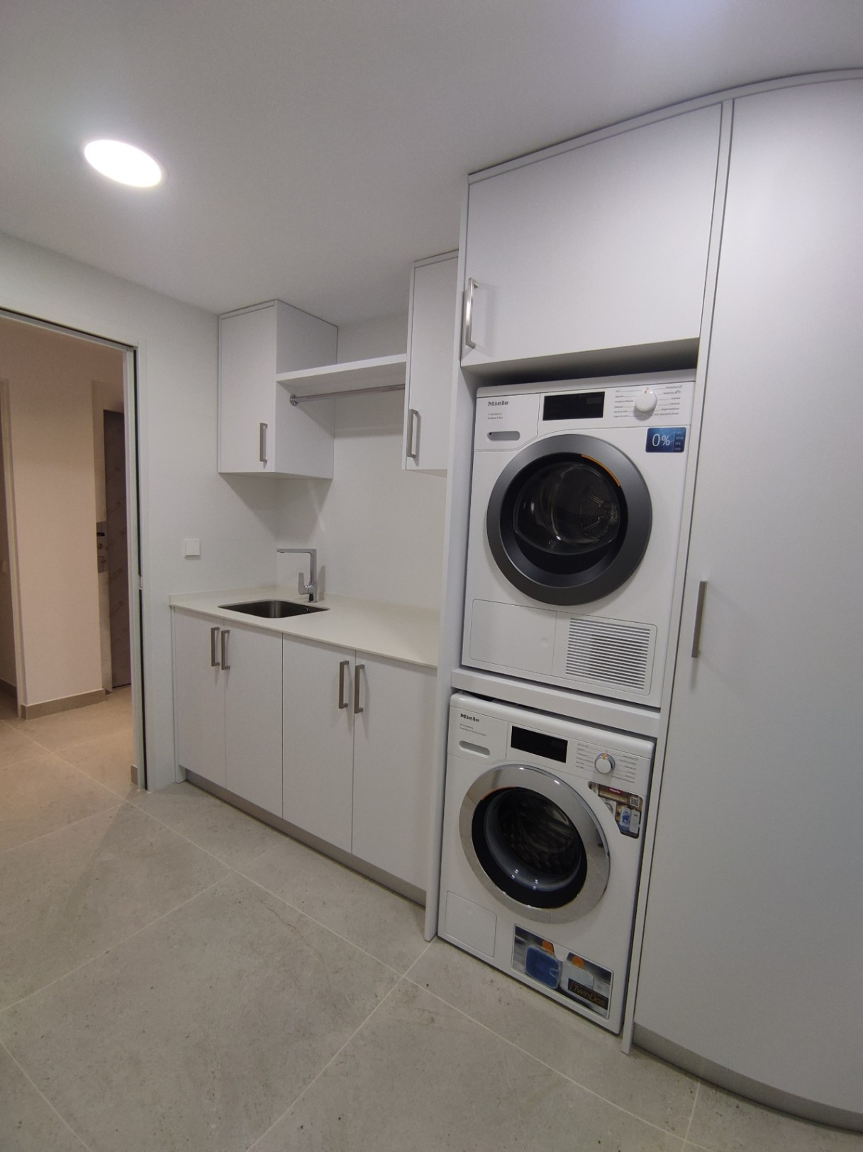 Functionele wasruimte met Miele-apparatuur in kolomopstelling, minimalistische witte kasten, geïntegreerde spoelbak en neutrale grootformaat vloertegels.