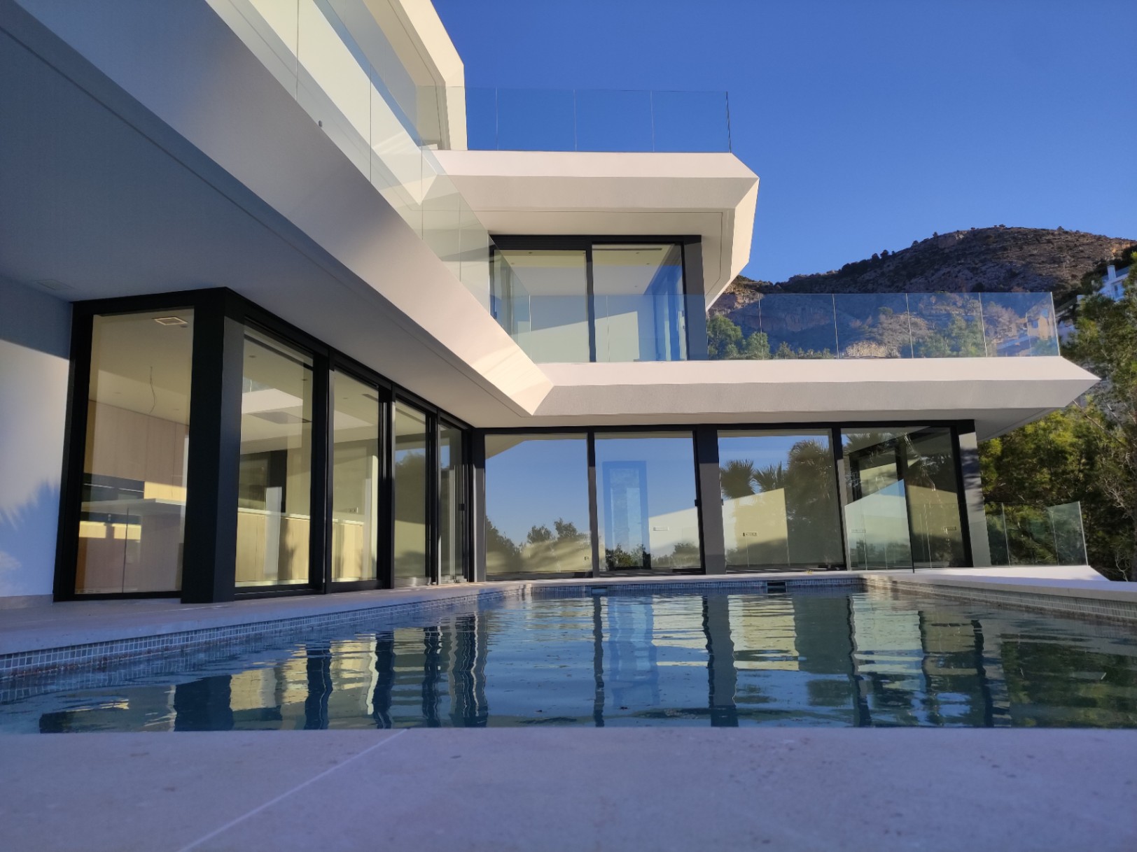 Moderne gevel met kamerhoge ramen in zwarte kozijnen en een overloopzwembad dat de geometrische architectuur weerspiegelt tegen de achtergrond van Altea Hills.