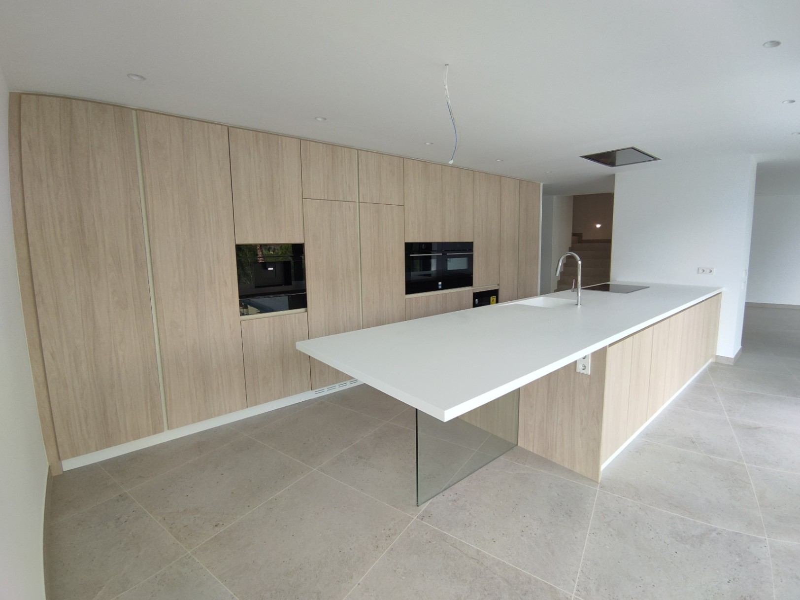 Moderne keuken met houten kastenwand, geïntegreerde apparatuur en een wit kookeiland met een glazen steunpoot en een neutrale tegelvloer.