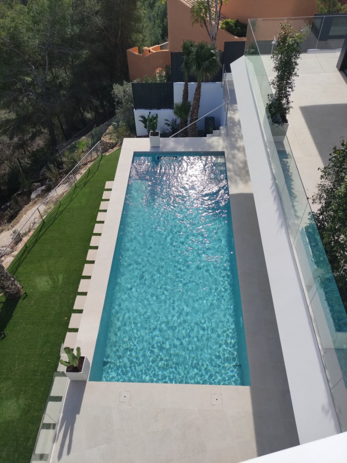 Rechthoekig zwembad met helderblauw water, omgeven door een licht stenen terras, kunstgras met stapstenen en glazen balustrades voor een modern design.