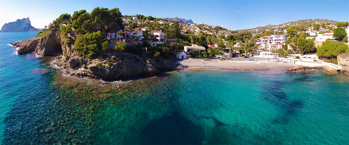 Luchtfoto van het strand van La Fustera met turquoise water, rotsachtige kliffen en de Peñón de Ifach in de verte, omgeven door pijnbomen en kustvilla's.