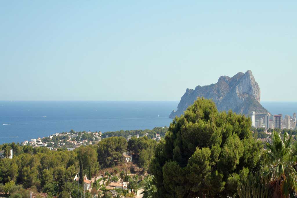 Panoramisch uitzicht op de Peñón de Ifach en de kustlijn van Calpe, met mediterrane dennenbossen en de blauwe zee onder een heldere hemel.