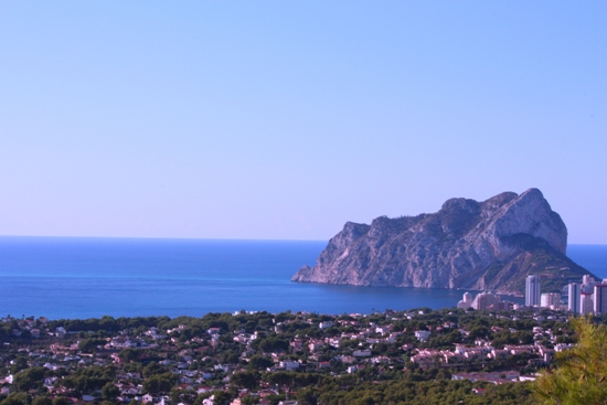 Panoramisch uitzicht op de Peñón de Ifach en de Middellandse Zee, met de kustlijn van Calpe en weelderig groen onder een helderblauwe hemel.