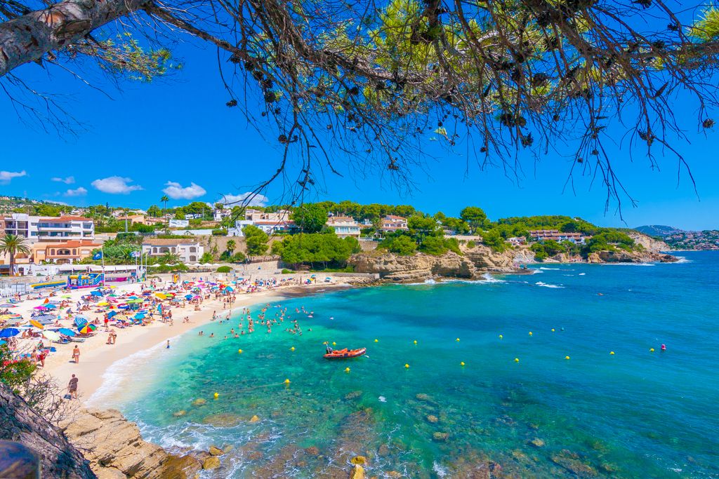 Panoramisch uitzicht op het zandstrand van La Fustera met turquoise water, omlijst door mediterrane pijnbomen en rotsachtige kustlijnen.