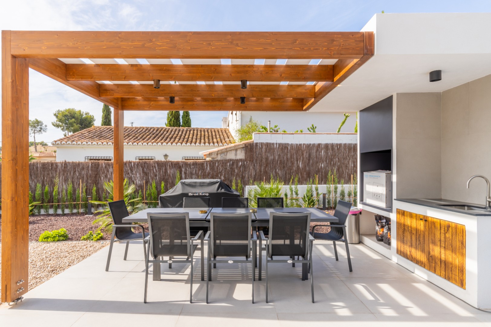 Detail van de eethoek buiten met een houten pergola en een buitenkeuken met spoelbak en barbecue.