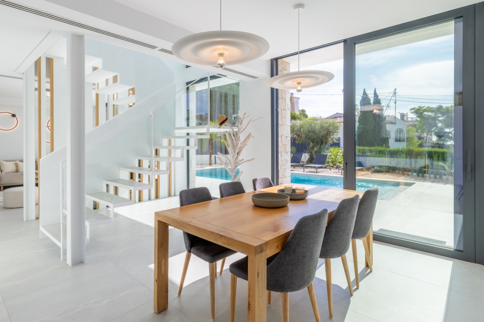 Lichte eetkamer met houten tafel, uitzicht op het zwembad en een witte zwevende trap.