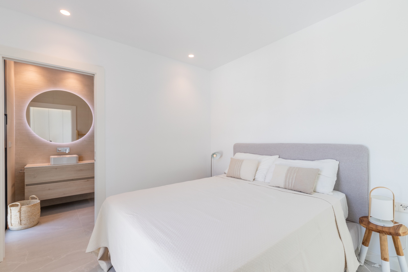 Lichte slaapkamer met ensuite badkamer. Tweepersoonsbed met grijs hoofdeinde en wit beddengoed. Badkamer met ronde verlichte spiegel.
