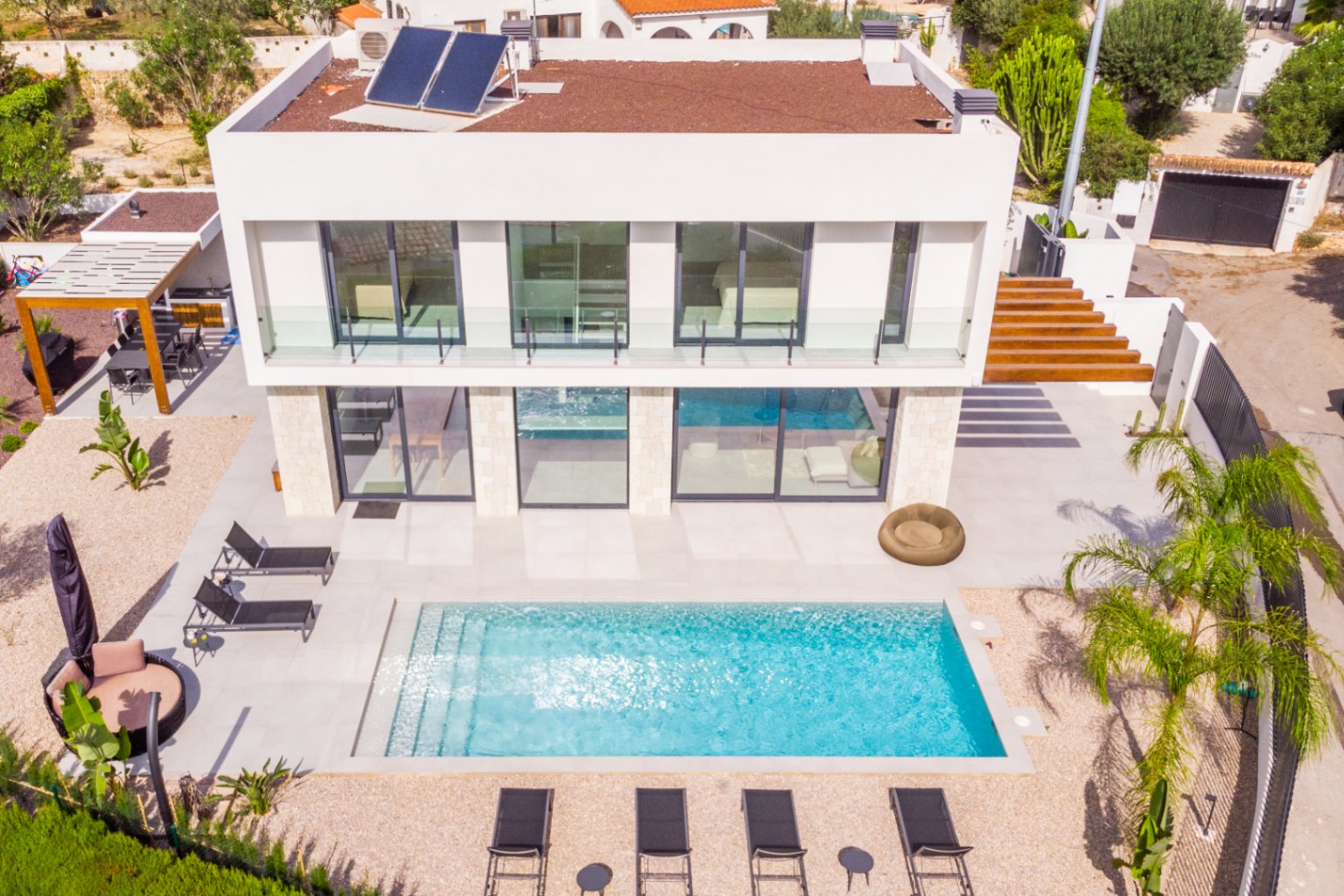 Luchtfoto van de villa met rechthoekig zwembad, omgeven door ligstoelen en licht grind. Modern design met zonnepanelen.