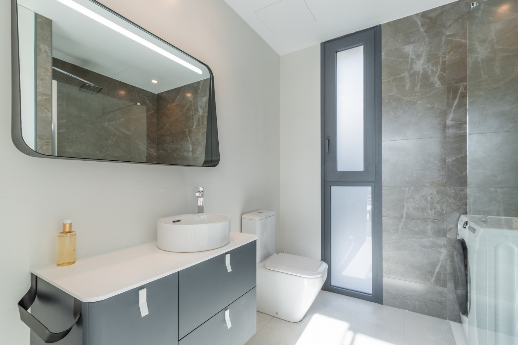 Modern badkamerdetail met keramische wastafel, spiegel met achtergrondverlichting en grijze marmeren bekleding.