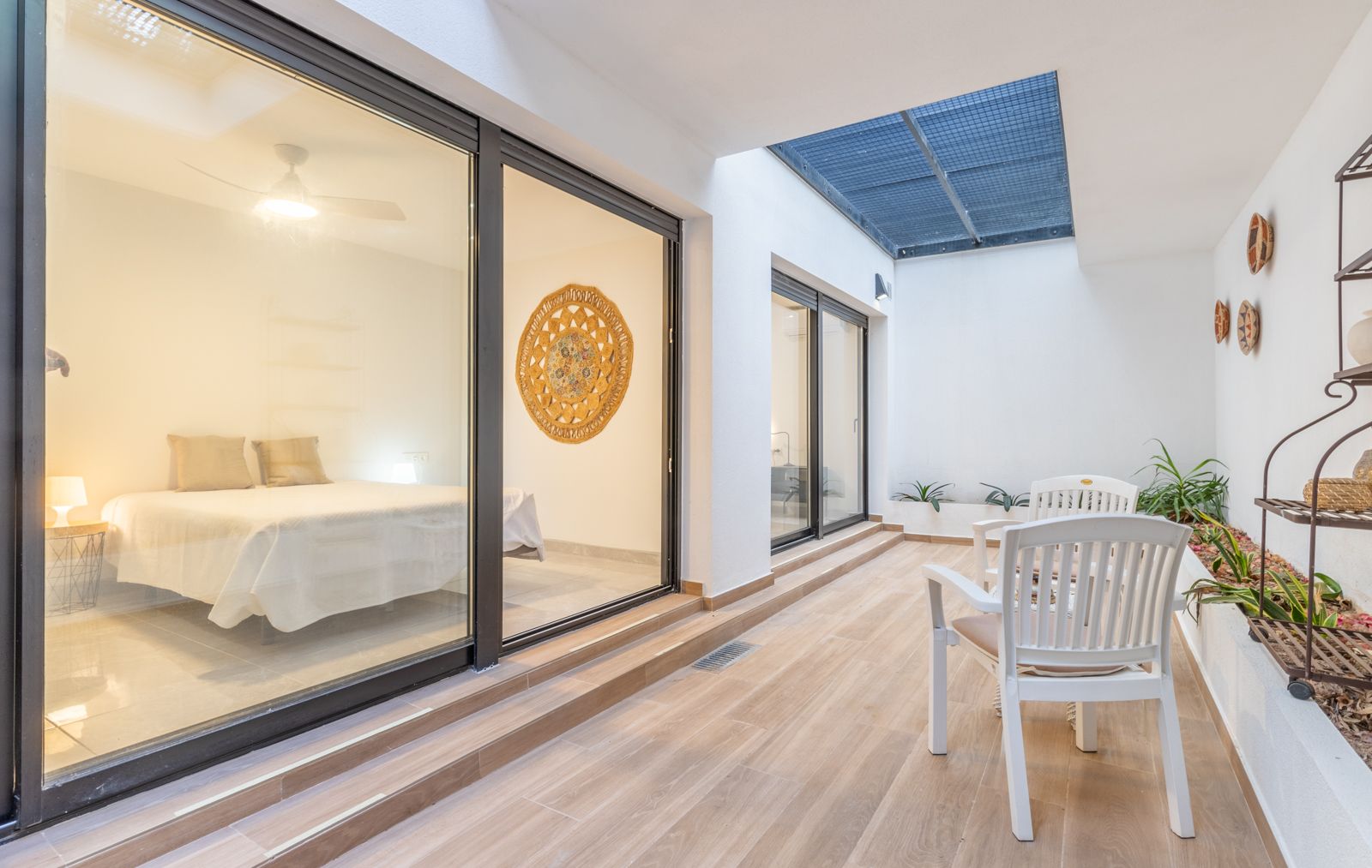 Binnenplaats met houtlook tegels, een zithoek en grote glazen schuifdeuren die toegang bieden tot een lichte slaapkamer met een strak design.