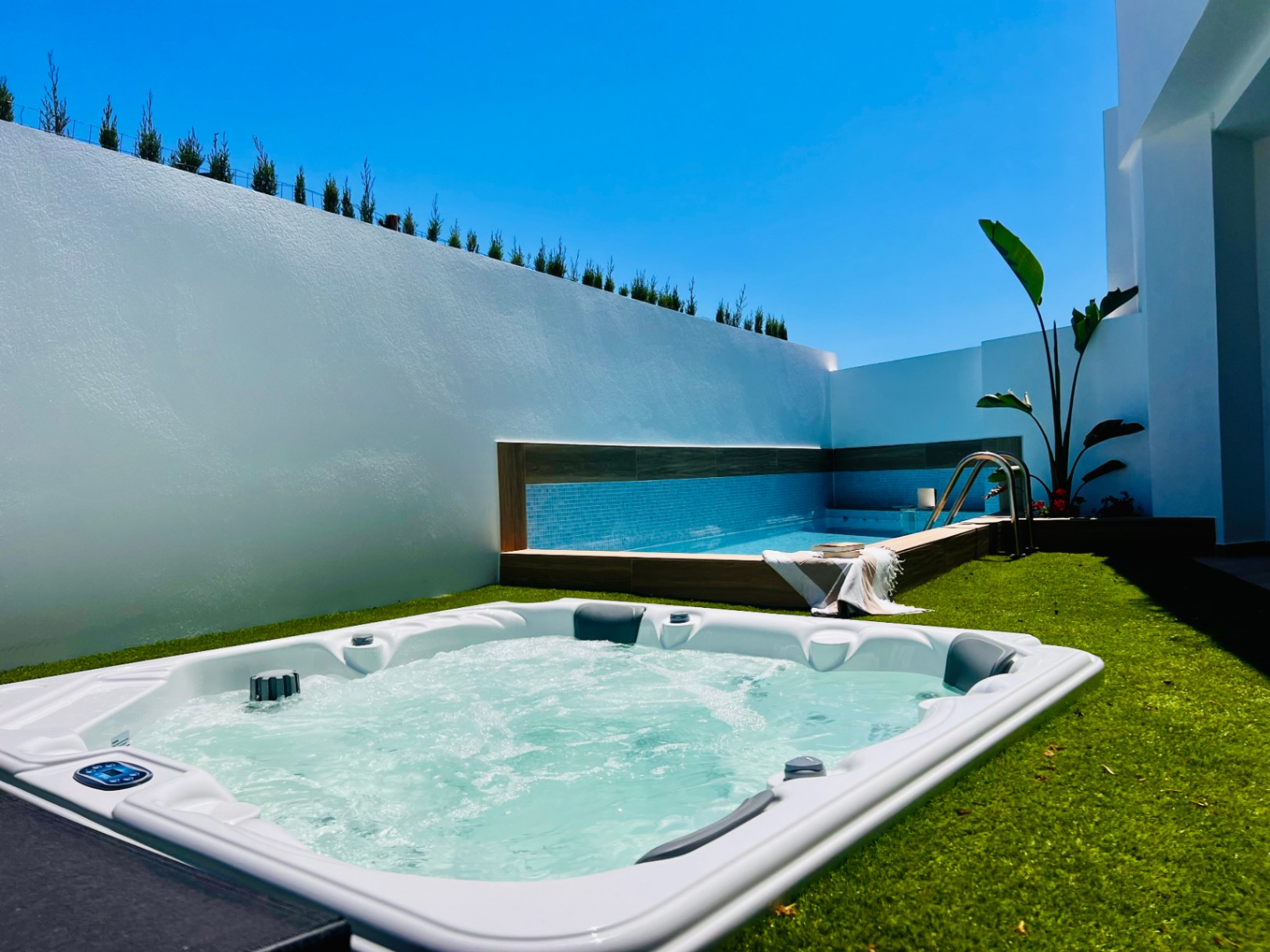 Buitenjacuzzi ingebed in kunstgras, gelegen naast een privézwembad met blauwe tegels en hoge witte muren voor optimale privacy.