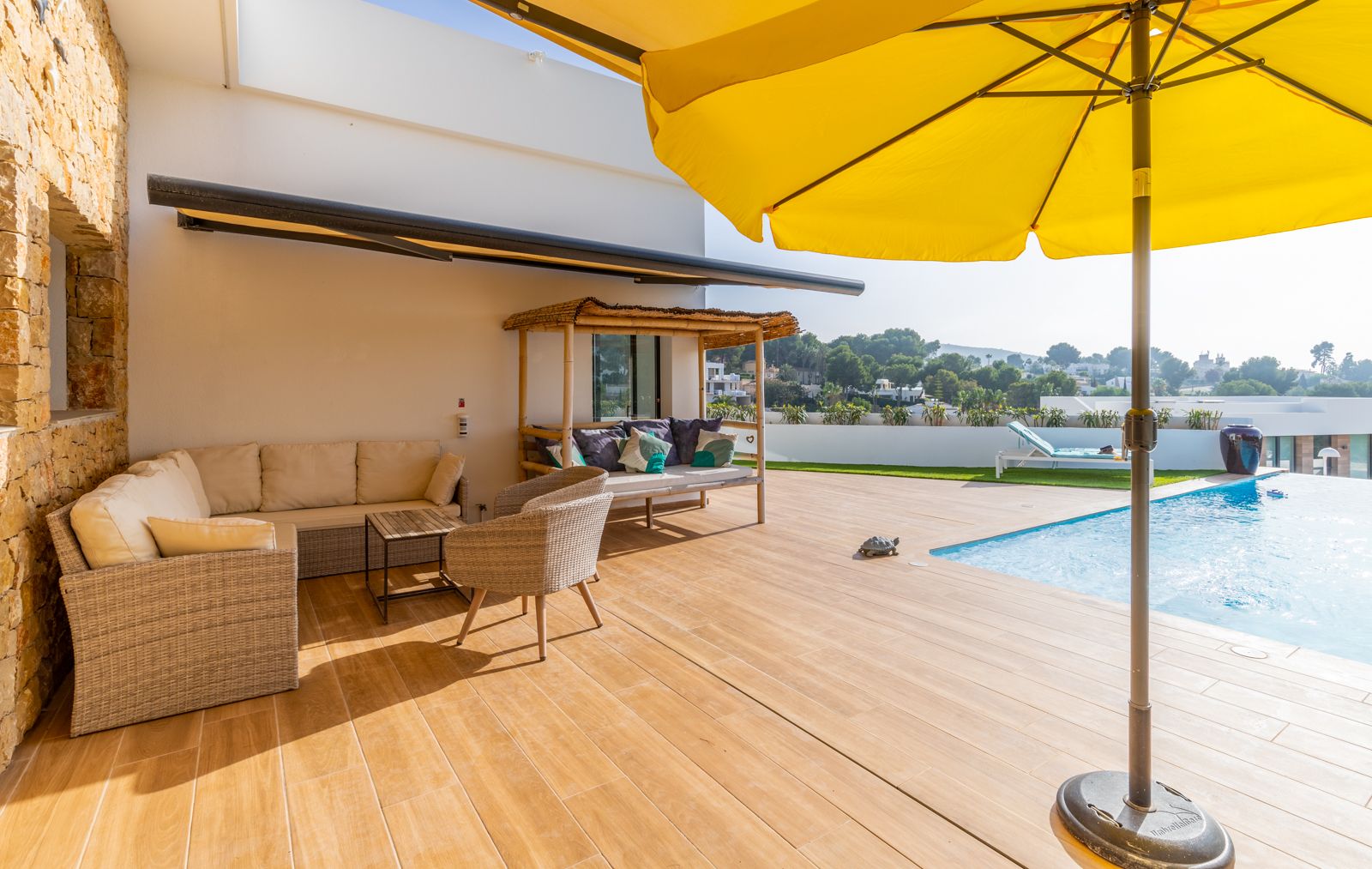 Buitenterras met houtlook keramische tegels, wicker loungeset, een bamboe dagbed en zwembad, afgewerkt met een natuurstenen muur en een gele parasol.