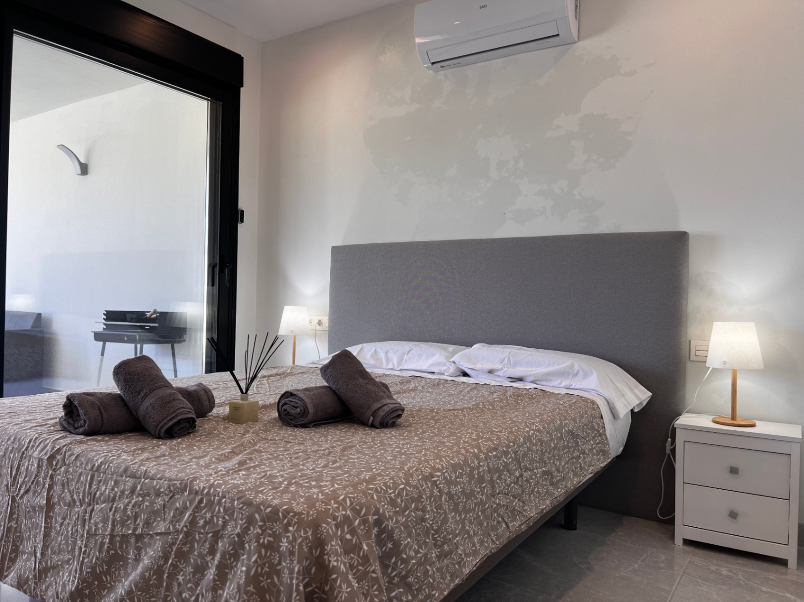 Eigentijdse slaapkamer met grijs stoffen hoofdbord, split-unit airconditioning en directe toegang tot het terras via grote glazen schuifdeuren met zwart kader.