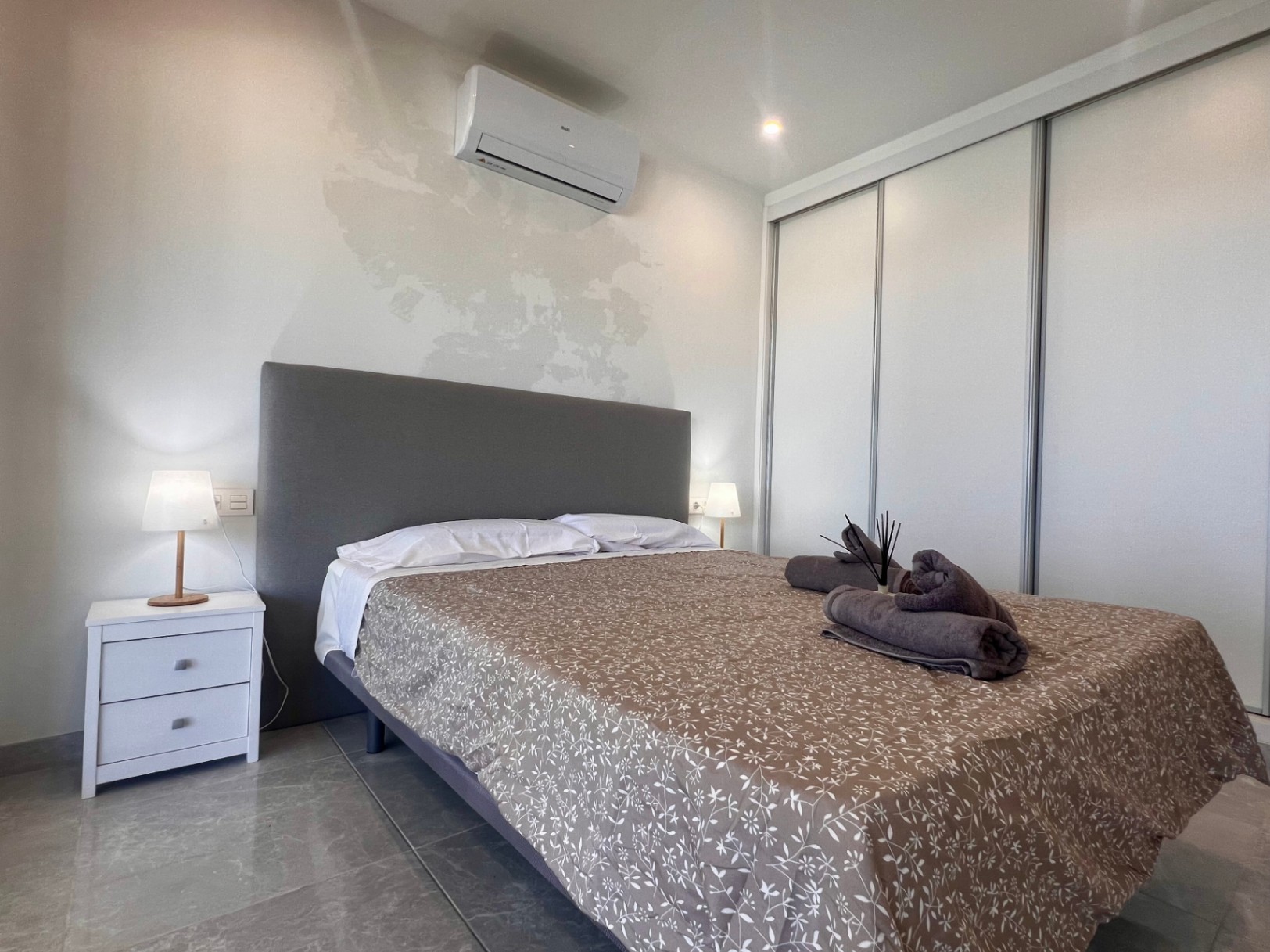 Eigentijdse slaapkamer met witte schuifdeurkast, airconditioning en een grijs gestoffeerd hoofdbord op een moderne tegelvloer.