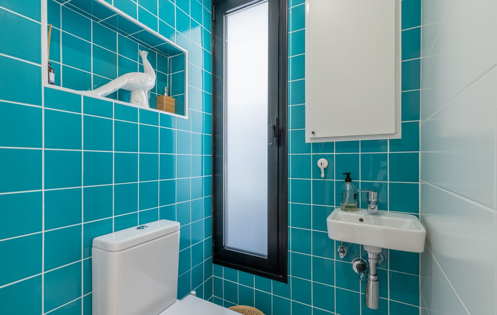 Gastentoilet met levendige turquoise tegels, een ingebouwde nis en een compacte wastafel. Een smal raam met melkglas zorgt voor natuurlijk licht en privacy.