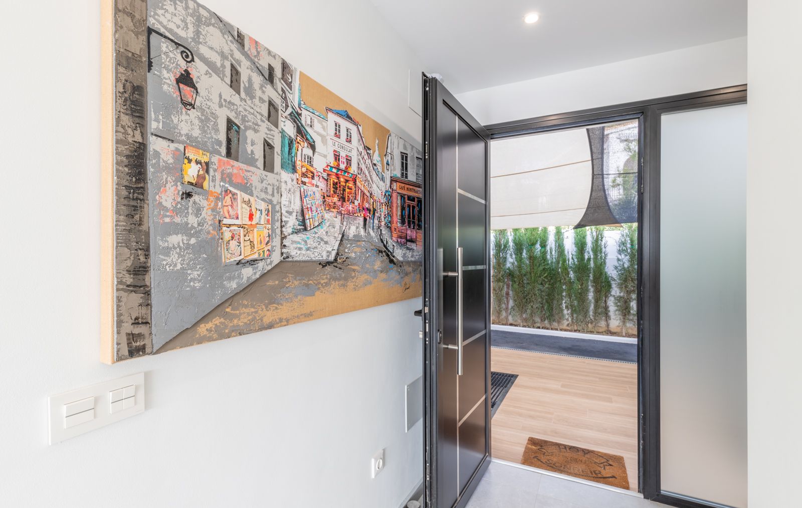 Hoofdentree met moderne antracietkleurige veiligheidsdeur, groot kunstwerk aan de wand en uitzicht op het houten terras met groen blijvende privacyhaag.