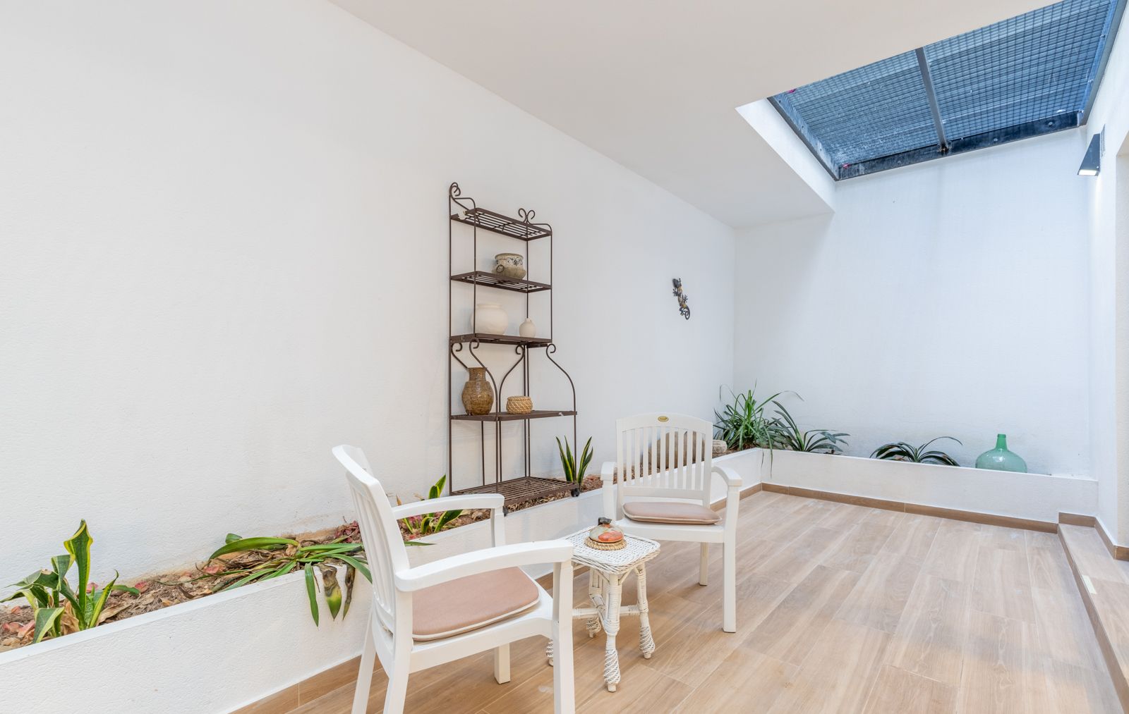 Lichte binnenplaats met dakraam, houtlook tegelvloer, geïntegreerde plantenbakken en een zithoek met witte stoelen en een smeedijzeren rek.