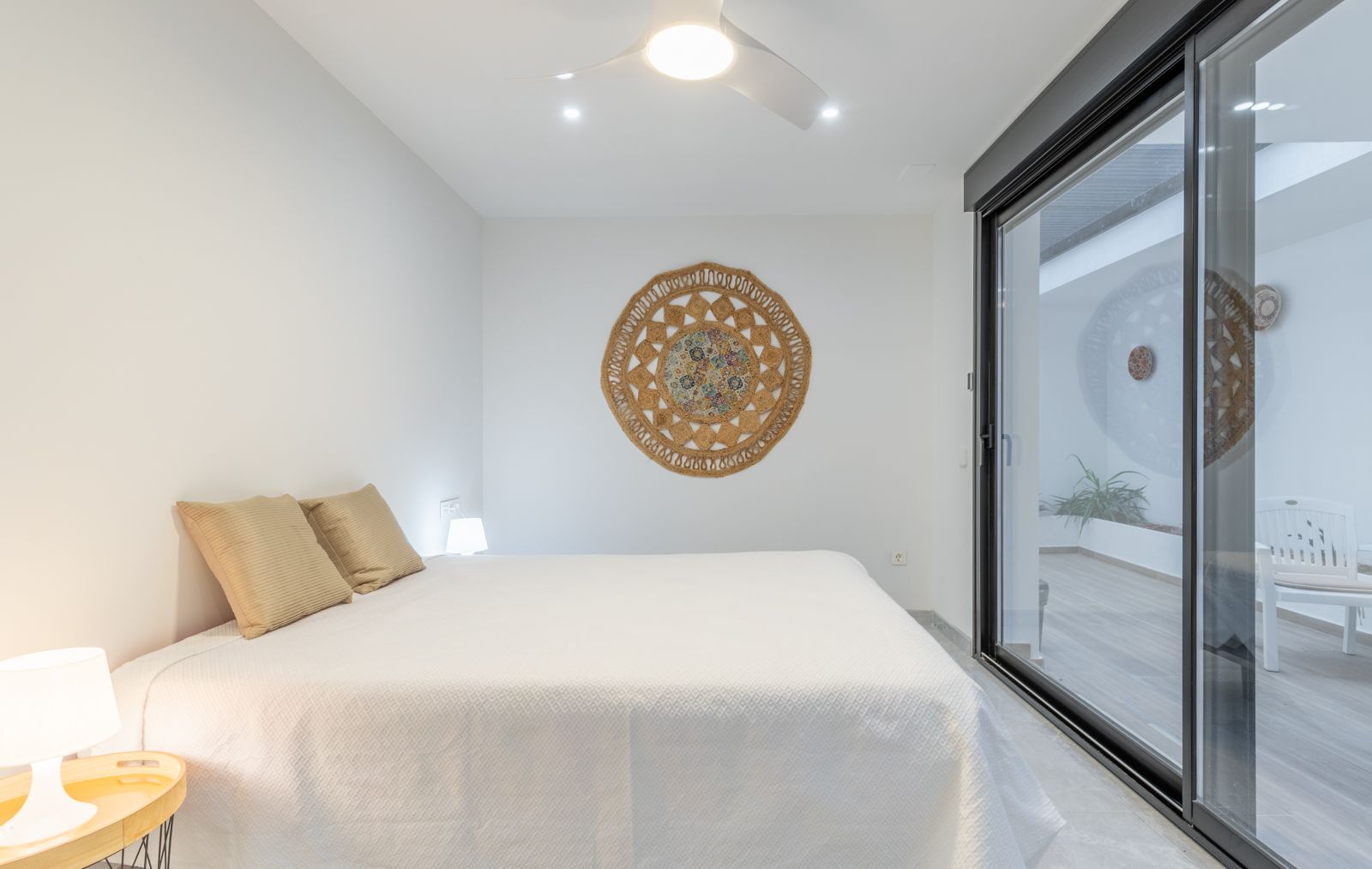 Minimalistische slaapkamer met directe toegang tot het terras via grote glazen schuifdeuren, voorzien van een plafondventilator en wanddecoratie van natuurlijke vezels.