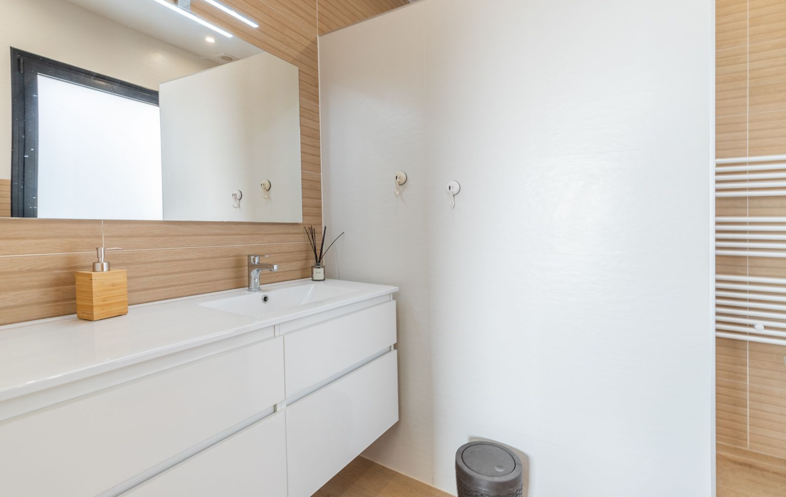 Moderne badkamer met een zwevend wit wastafelmeubel, wandtegels met houtlook, een grote spiegel met LED-verlichting en een witte handdoekradiator.