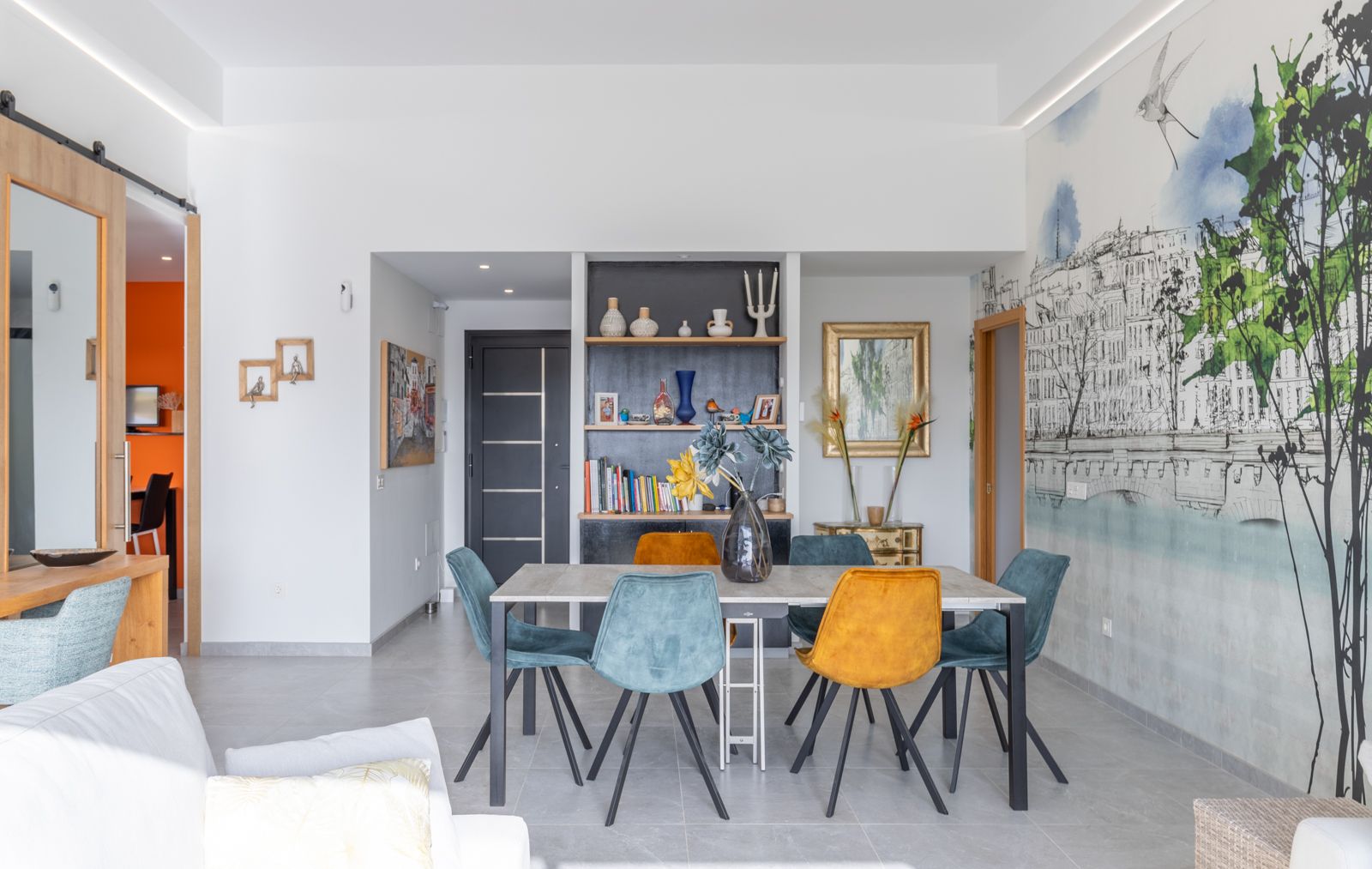 Moderne eethoek met een minimalistische tafel en fluwelen stoelen in blauw en oker. De ruimte is voorzien van een artistieke muurschildering en ingebouwde kasten met LED-verlichting.