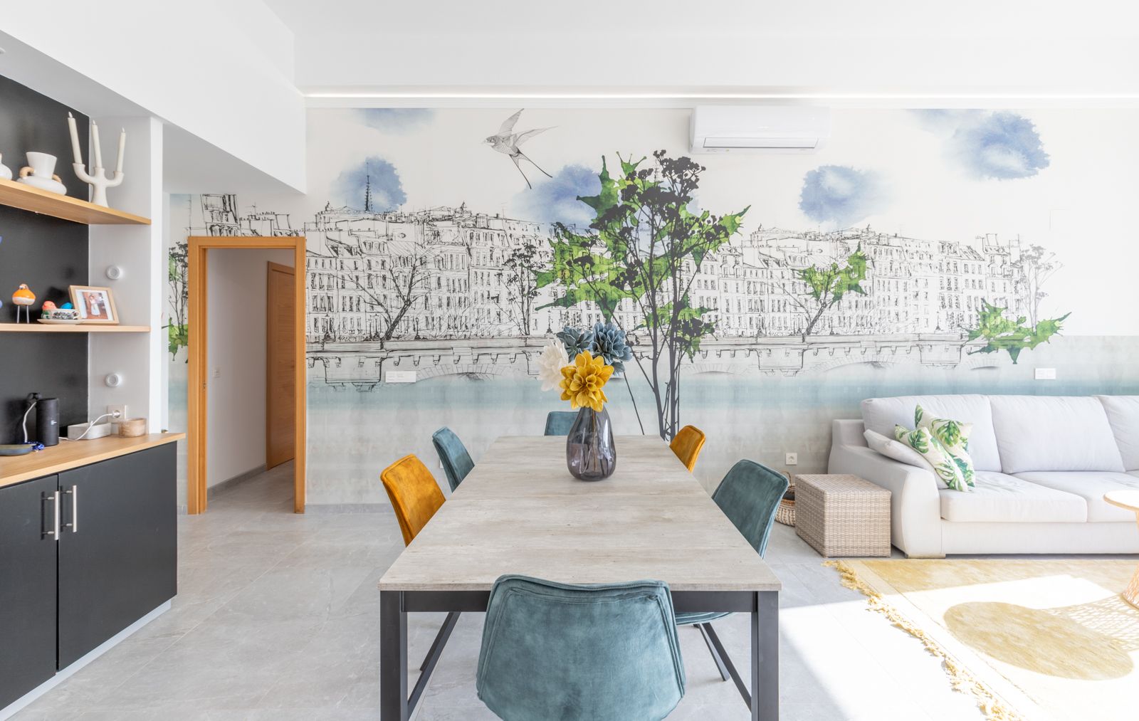 Moderne eethoek met tafel in steenlook, fluwelen stoelen in blauw en goudgeel, en een grote muurschildering. Grijze tegelvloer en aangrenzende zithoek.