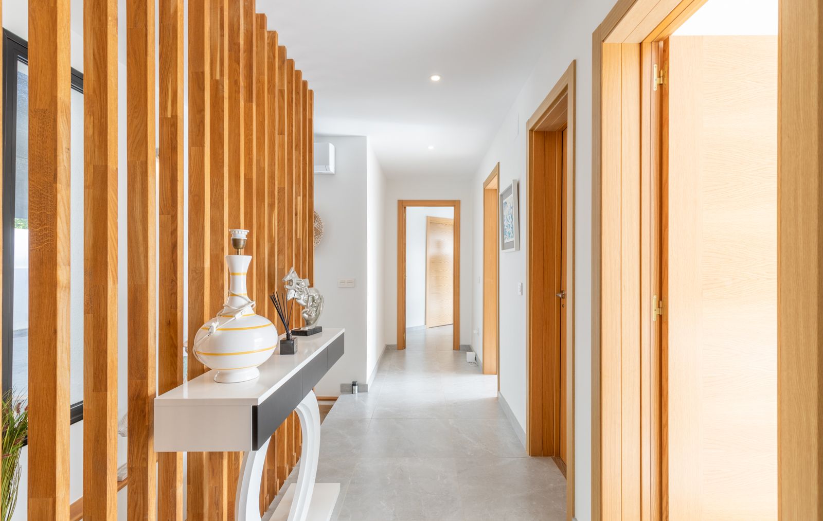 Moderne gang met een verticale houten lattenwand, grijze keramische vloertegels en bijpassende licht houten deurkozijnen.