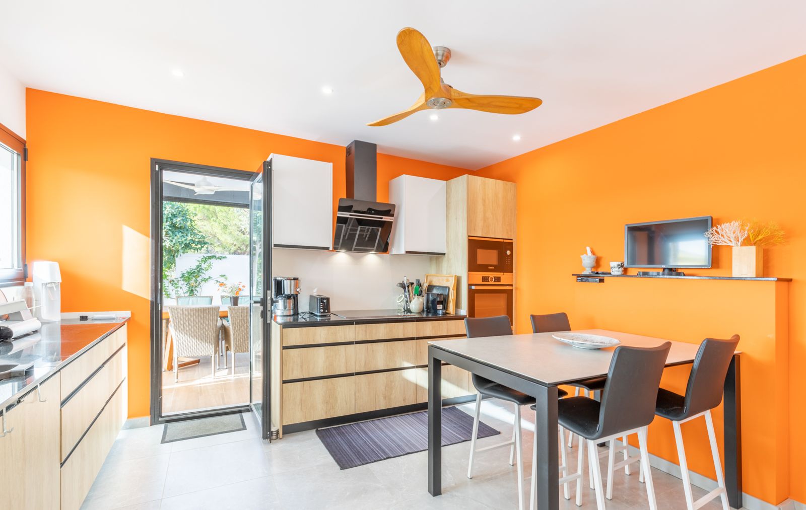 Moderne keuken met levendige oranje muren, lichte houten kasten en een geïntegreerde eethoek met directe toegang tot het buitenterras.