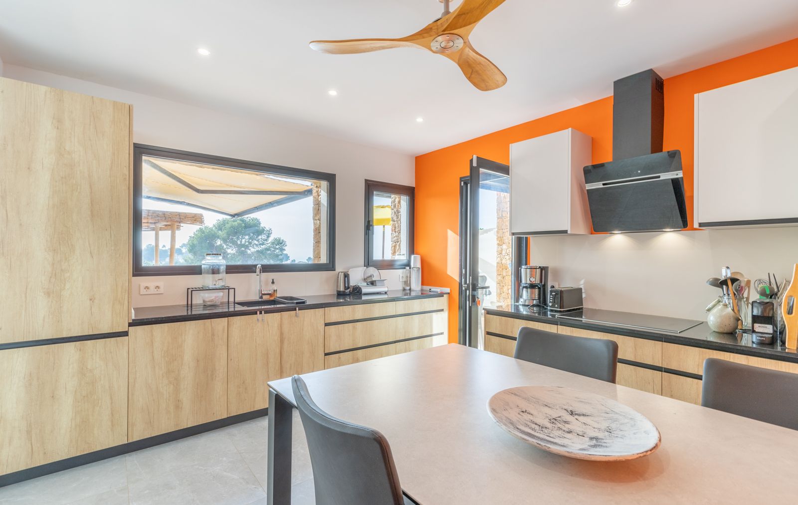 Moderne keuken met lichte houten kasten, zwarte werkbladen en een opvallende oranje wand. Voorzien van een houten plafondventilator en grote ramen met zicht op het terras.